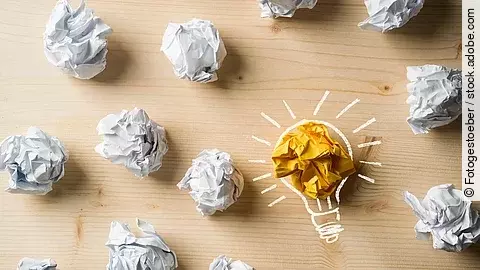 Viele zusammengeknüllte Blätter Papier. Eines davon in gelber Farbe und umrahmt wie eine leuchtende Glühbirne.