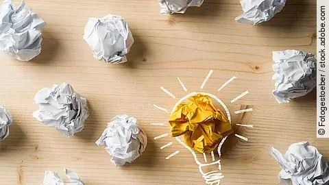 Viele zusammengeknüllte Blätter Papier. Eines davon in gelber Farbe und umrahmt wie eine leuchtende Glühbirne.