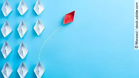 Change Management Mehrere gefaltete weiße Papierschiffchen sind von oben zu sehen. Ein rotes Papierschiffchen schwimmt heraus aus der Formation.