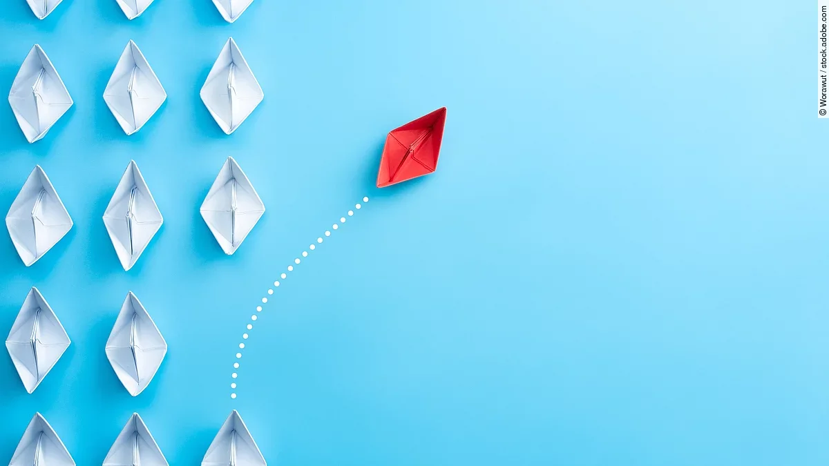 Change Management Mehrere gefaltete weiße Papierschiffchen sind von oben zu sehen. Ein rotes Papierschiffchen schwimmt heraus aus der Formation.