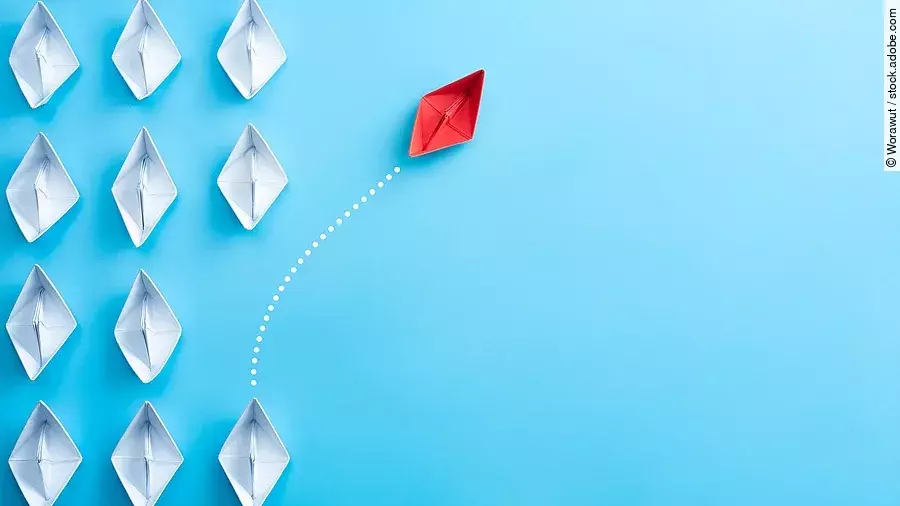 Mehrere gefaltete weiße Papierschiffchen sind von oben zu sehen. Ein rotes Papierschiffchen schwimmt heraus aus der Formation.
