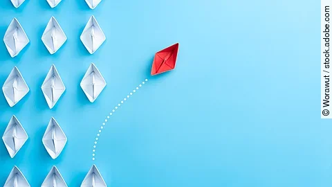 Mehrere gefaltete weiße Papierschiffchen sind von oben zu sehen. Ein rotes Papierschiffchen schwimmt heraus aus der Formation.