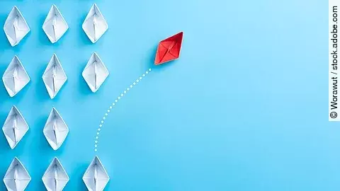 Mehrere gefaltete weiße Papierschiffchen sind von oben zu sehen. Ein rotes Papierschiffchen schwimmt heraus aus der Formation.