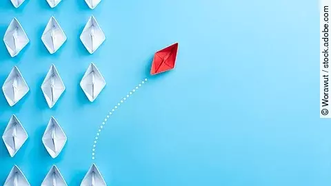 Mehrere gefaltete weiße Papierschiffchen sind von oben zu sehen. Ein rotes Papierschiffchen schwimmt heraus aus der Formation.