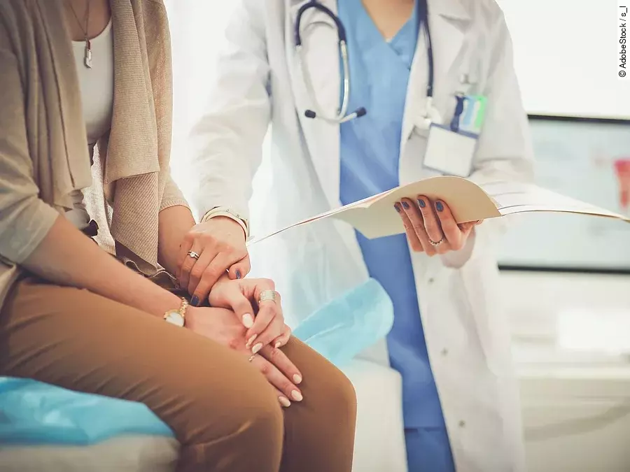 Ein Arzt greift den Arm seiner Patientin und hält die Patientenakte in der anderen Hand. Im Hintergrund steht ein Bildschirm.