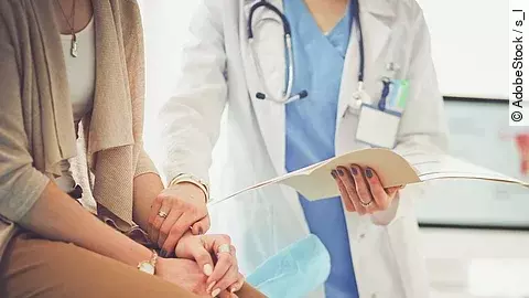 Ein Arzt greift den Arm seiner Patientin und hält die Patientenakte in der anderen Hand. Im Hintergrund steht ein Bildschirm.