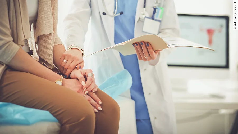 Ein Arzt greift den Arm seiner Patientin und hält die Patientenakte in der anderen Hand. Im Hintergrund steht ein Bildschirm.