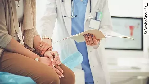 Ein Arzt greift den Arm seiner Patientin und hält die Patientenakte in der anderen Hand. Im Hintergrund steht ein Bildschirm.