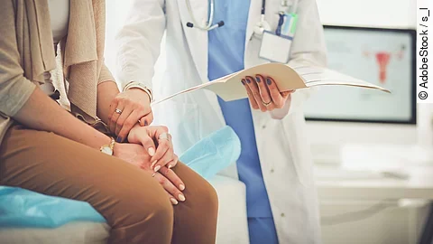 Ein Arzt greift den Arm seiner Patientin und hält die Patientenakte in der anderen Hand. Im Hintergrund steht ein Bildschirm.