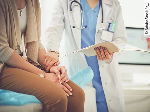 Ein Arzt greift den Arm seiner Patientin und hält die Patientenakte in der anderen Hand. Im Hintergrund steht ein Bildschirm.