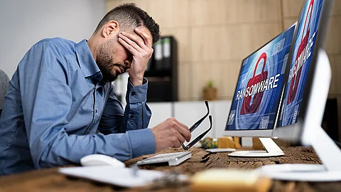 Ein Mann sitzt am Schreibtisch und verschränkt die Hand vorm Gesicht. Auf seinem Computerbildschirm steht das Wort "Ransomeware". Es erscheint der Eindruck, dass der Mann von einem Cyberangriff betroffen ist.