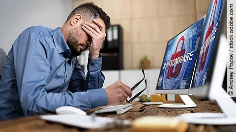 Ein Mann sitzt am Schreibtisch und verschränkt die Hand vorm Gesicht. Auf seinem Computerbildschirm steht das Wort "Ransomeware". Es erscheint der Eindruck, dass der Mann von einem Cyberangriff betroffen ist.