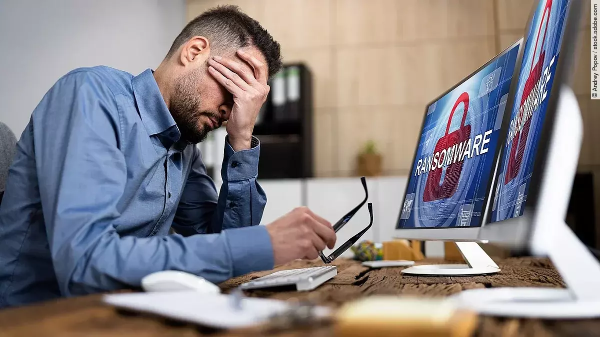 Ein Mann sitzt am Schreibtisch und verschränkt die Hand vorm Gesicht. Auf seinem Computerbildschirm steht das Wort "Ransomeware". Es erscheint der Eindruck, dass der Mann von einem Cyberangriff betroffen ist.