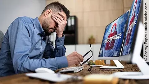 Ein Mann sitzt am Schreibtisch und verschränkt die Hand vorm Gesicht. Auf seinem Computerbildschirm steht das Wort "Ransomeware". Es erscheint der Eindruck, dass der Mann von einem Cyberangriff betroffen ist.