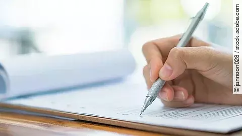 Eine Hand hält einen Stift und schreibt auf ein Prüfungsdokument.