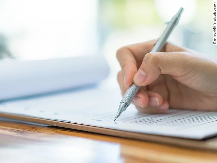 Eine Hand hält einen Stift und schreibt auf ein Prüfungsdokument.