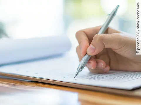 Eine Hand hält einen Stift und schreibt auf ein Prüfungsdokument.