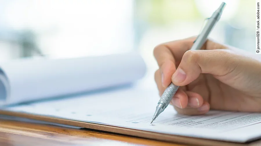 Eine Hand hält einen Stift und schreibt auf ein Prüfungsdokument.