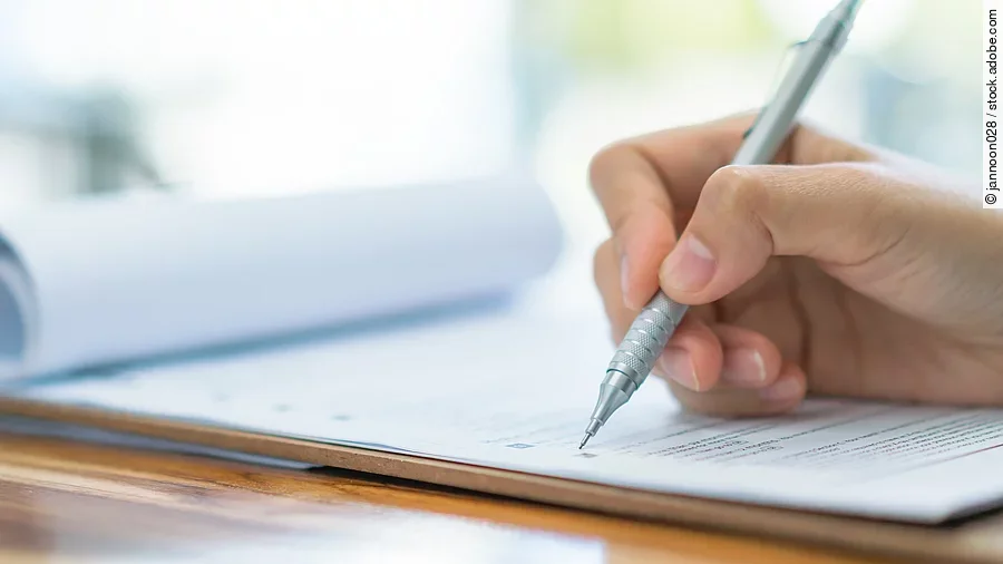 Eine Hand hält einen Stift und schreibt auf ein Prüfungsdokument.