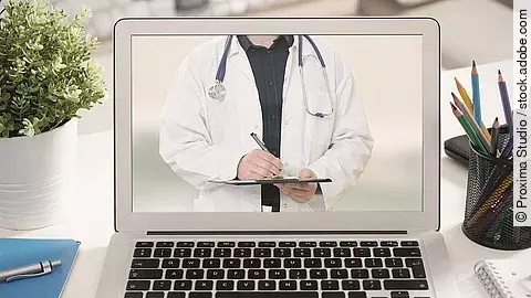 Telemedizin Ein aufgeklappter Laptop steht auf einem Tisch. Der Bildschirm zeigt den Torso eines Arztes.