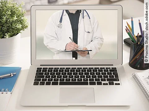 Ein aufgeklappter Laptop steht auf einem Tisch. Der Bildschirm zeigt den Torso eines Arztes.