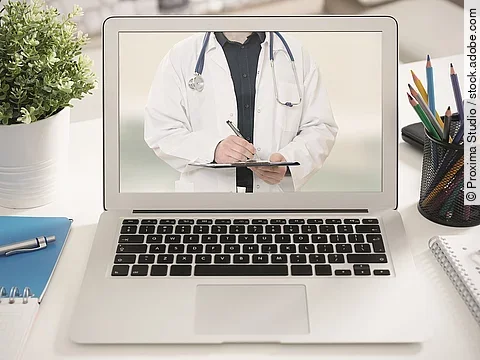 Ein aufgeklappter Laptop steht auf einem Tisch. Der Bildschirm zeigt den Torso eines Arztes.