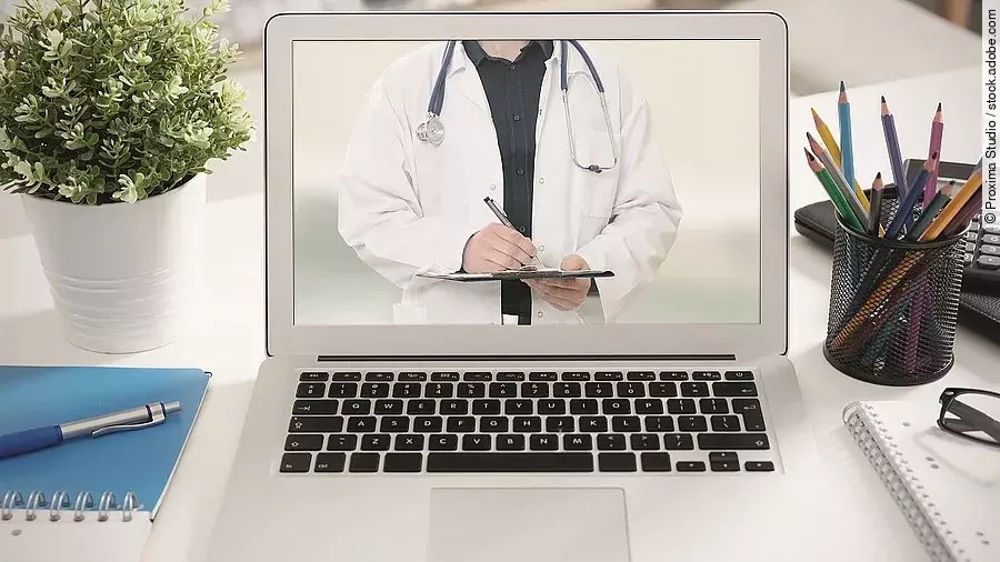 Ein aufgeklappter Laptop steht auf einem Tisch. Der Bildschirm zeigt den Torso eines Arztes.
