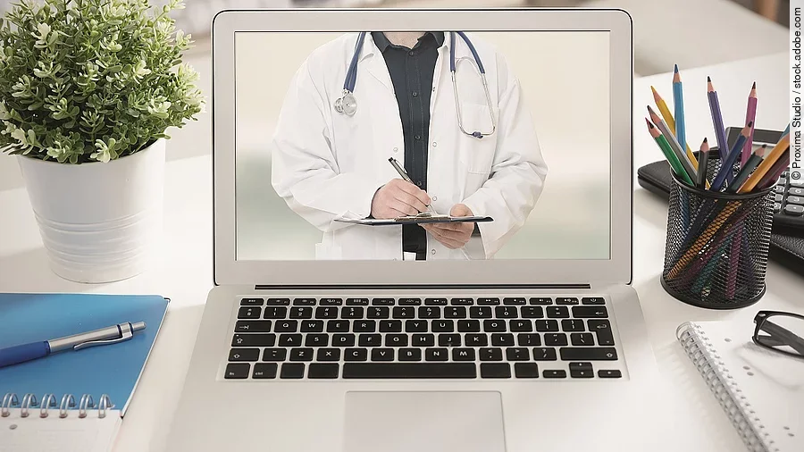 Telemedizin Ein aufgeklappter Laptop steht auf einem Tisch. Der Bildschirm zeigt den Torso eines Arztes.
