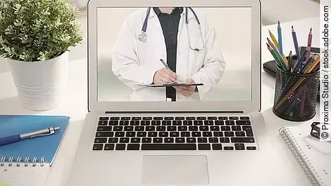 Ein aufgeklappter Laptop steht auf einem Tisch. Der Bildschirm zeigt den Torso eines Arztes.