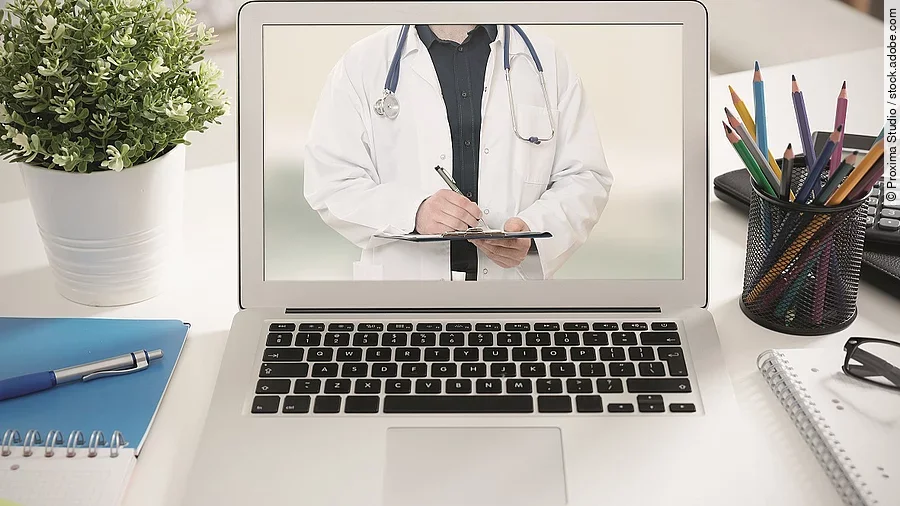 Ein aufgeklappter Laptop steht auf einem Tisch. Der Bildschirm zeigt den Torso eines Arztes.