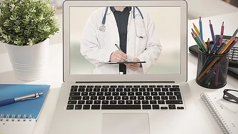 Ein aufgeklappter Laptop steht auf einem Tisch. Der Bildschirm zeigt den Torso eines Arztes.