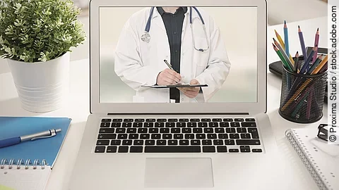 Telemedizin Ein aufgeklappter Laptop steht auf einem Tisch. Der Bildschirm zeigt den Torso eines Arztes.