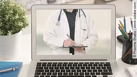 Ein aufgeklappter Laptop steht auf einem Tisch. Der Bildschirm zeigt den Torso eines Arztes.