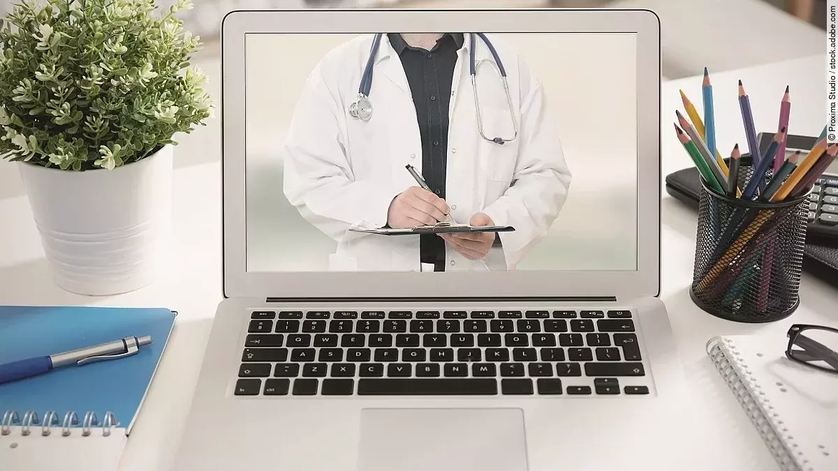 Telemedizin Ein aufgeklappter Laptop steht auf einem Tisch. Der Bildschirm zeigt den Torso eines Arztes.