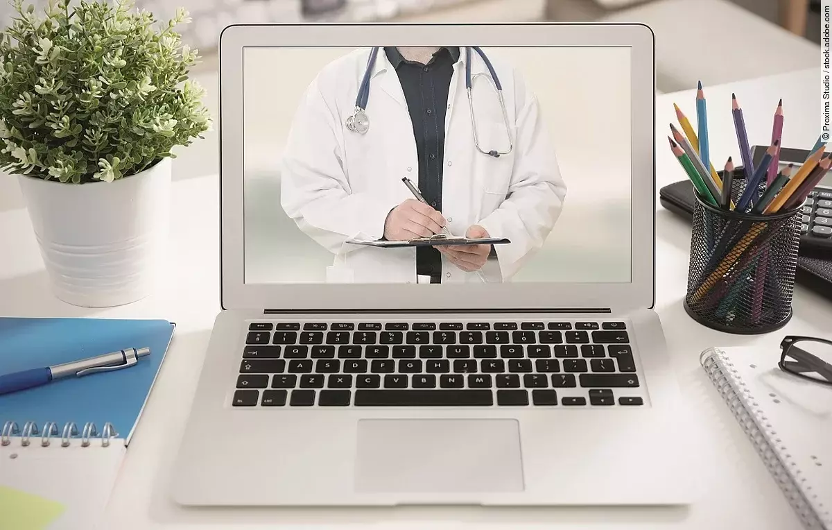 Telemedizin Ein aufgeklappter Laptop steht auf einem Tisch. Der Bildschirm zeigt den Torso eines Arztes.