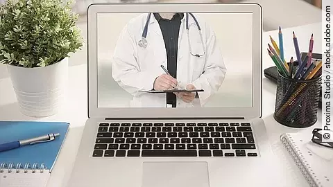 Telemedizin Ein aufgeklappter Laptop steht auf einem Tisch. Der Bildschirm zeigt den Torso eines Arztes.