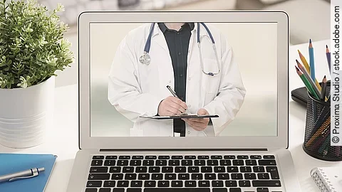 Ein aufgeklappter Laptop steht auf einem Tisch. Der Bildschirm zeigt den Torso eines Arztes.
