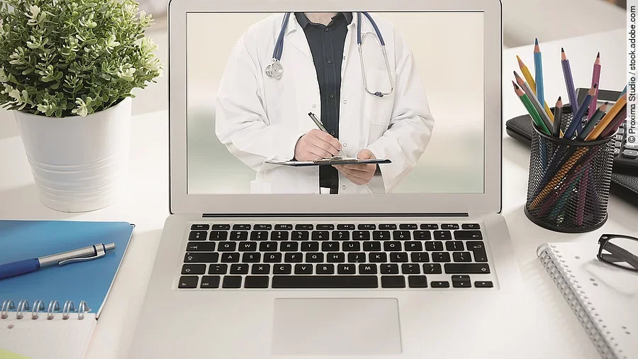 Ein aufgeklappter Laptop steht auf einem Tisch. Der Bildschirm zeigt den Torso eines Arztes.