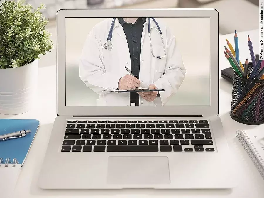 Ein aufgeklappter Laptop steht auf einem Tisch. Der Bildschirm zeigt den Torso eines Arztes.