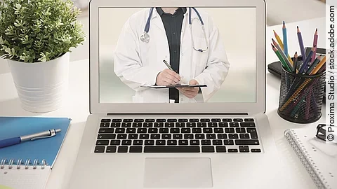 Ein aufgeklappter Laptop steht auf einem Tisch. Der Bildschirm zeigt den Torso eines Arztes.