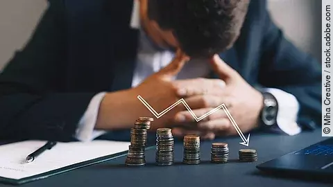 Insolvenz Ein Mann im Anzug, der mit gesenktem Kopf und verschränkten Händen vor sich sitzt. Auf dem Tisch vor ihm liegen gestapelte Münzen, über ihnen ist eine abwärtsführende Diagrammlinie eingezeichnet.