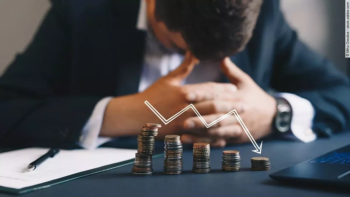 Ein Mann im Anzug, der mit gesenktem Kopf und verschränkten Händen vor sich sitzt. Auf dem Tisch vor ihm liegen gestapelte Münzen, über ihnen ist eine abwärtsführende Diagrammlinie eingezeichnet. 