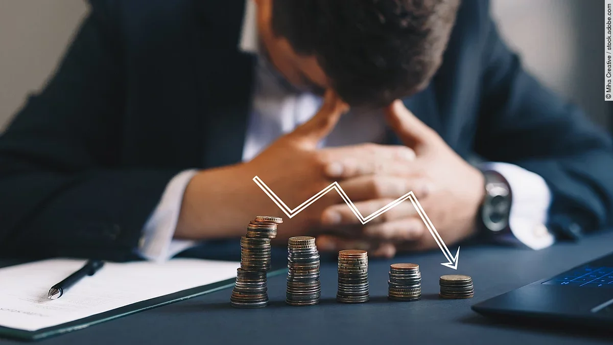 Ein Mann im Anzug, der mit gesenktem Kopf und verschränkten Händen vor sich sitzt. Auf dem Tisch vor ihm liegen gestapelte Münzen, über ihnen ist eine abwärtsführende Diagrammlinie eingezeichnet. 