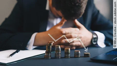 Ein Mann im Anzug, der mit gesenktem Kopf und verschränkten Händen vor sich sitzt. Auf dem Tisch vor ihm liegen gestapelte Münzen, über ihnen ist eine abwärtsführende Diagrammlinie eingezeichnet. 