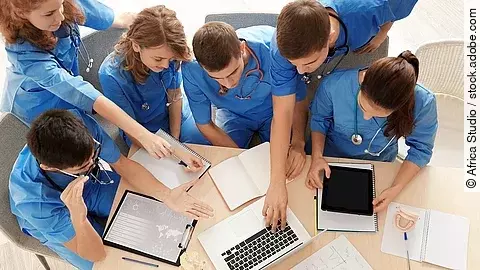 Aus der Vogelperspektive sind sechs Medizinstudenten in blauen Kasacks zu sehen, sie lernen zusammen und sprechen über etwas, das auf einem Laptop zusehen ist. 