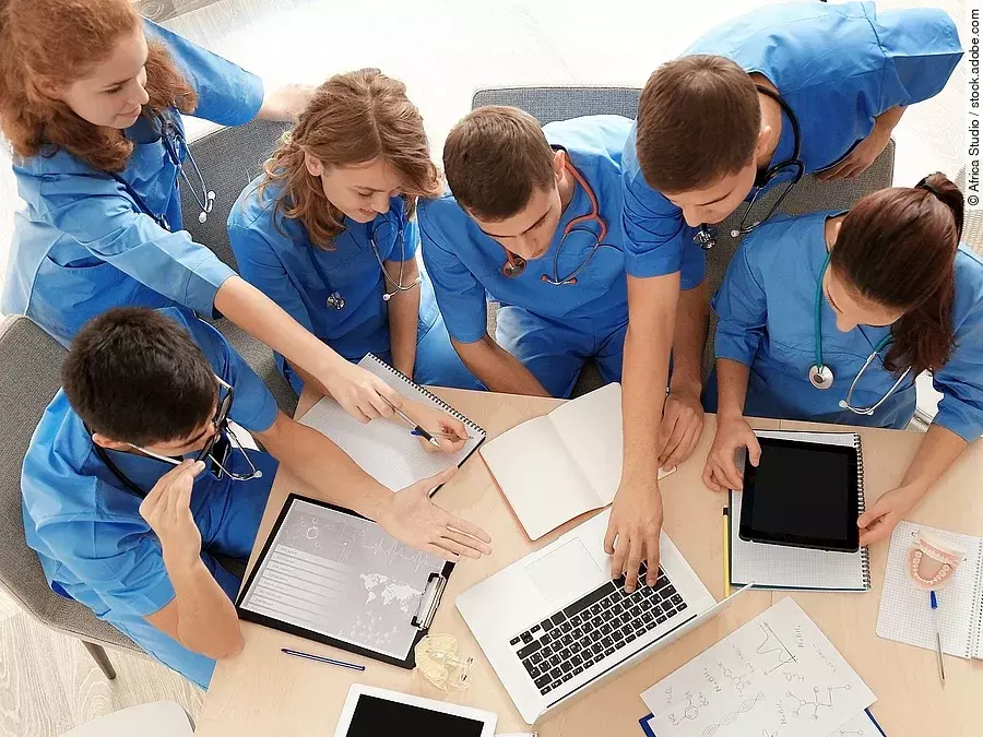 Aus der Vogelperspektive sind sechs Medizinstudenten in blauen Kasacks zu sehen, sie lernen zusammen und sprechen über etwas, das auf einem Laptop zusehen ist. 