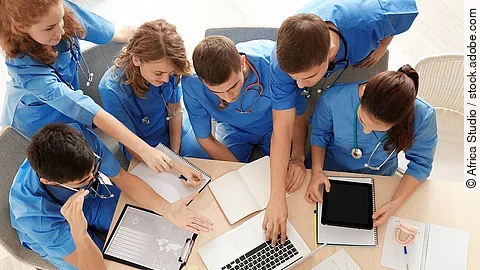 Medizinstudenten Aus der Vogelperspektive sind sechs Medizinstudenten in blauen Kasacks zu sehen, sie lernen zusammen und sprechen über etwas, das auf einem Laptop zusehen ist.