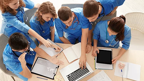 Medizinstudenten Aus der Vogelperspektive sind sechs Medizinstudenten in blauen Kasacks zu sehen, sie lernen zusammen und sprechen über etwas, das auf einem Laptop zusehen ist.