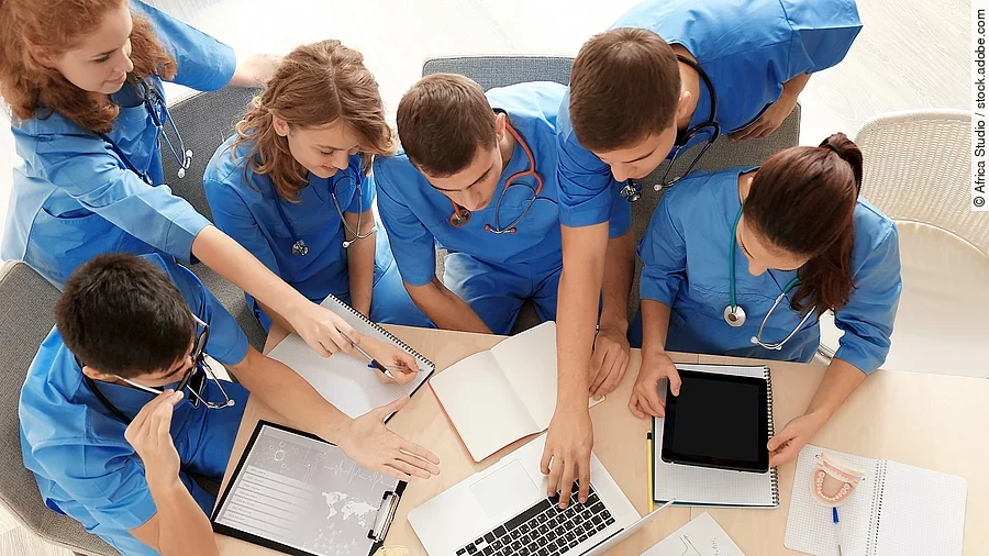 Aus der Vogelperspektive sind sechs Medizinstudenten in blauen Kasacks zu sehen, sie lernen zusammen und sprechen über etwas, das auf einem Laptop zusehen ist. 