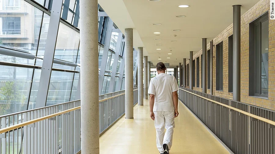Mensch in weißer Arbeitskleidung läuft, den Rückend zugewandt, einen Krankenhausflur entlang.